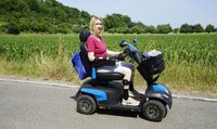 Eine Frau mit blonden Haaren sitzt in einem Elektro-Rollstuhl vor einem dicht bewachsenen Feld. Ihr Gesichtsausdruck ist ernst. 