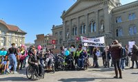 Eine Gruppe Menschen mit und ohne Beeinträchtigung ist auf einem Platz versammelt und hält Protestschilder hoch.