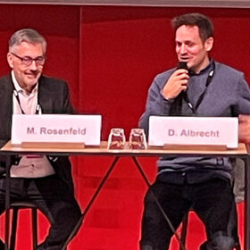 Martin D. Rosenfeld und Daniel Albrecht sitzen am Podium. Daniel Albrecht hält ein Mikrofon in der Hand.