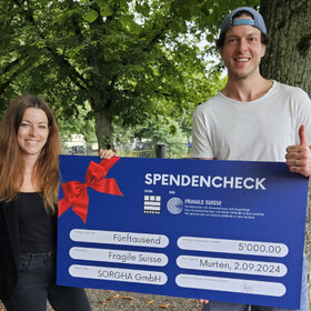 Zwei Personen posieren mit einem grossen blauen Spendencheck: links Jana Bauer, rechts Kevin Lötscher. Sie lächeln und halten den Daumen hoch.