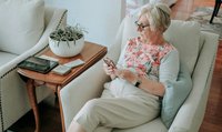 Une femme est assise dans son salon sur un fauteuil, les yeux rivés sur son téléphone portable.