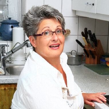 Portraitfoto einer Frau mit Brille und grauen Haaren. Im Hintergrund ist eine Küche.