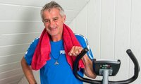 Portrait d'un homme debout à côté de son home-trainer. Il porte un t-shirt de sport bleu et un foulard rouge autour de la nuque.