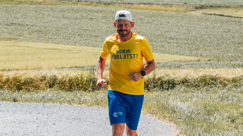 Iwan B. Ein Mann läuft auf einer Landstrasse. Er trägt ein weisses Cap, ein gelbes FRAGILE-T-Shirt und blaue kurze Hosen mit dem FRAGILE-Logo.