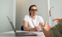 Un jeune homme portant des lunettes de soleil écoute une personne en face de lui. À côté de lui se trouvent un ordinateur et une canne d'aveugle.