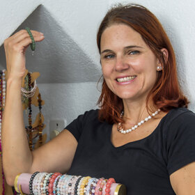  Claudia P, une femme aux cheveux roux qui lui arrivent aux épaules, sourit à la caméra et tient en l'air un bracelet de perles vertes. Dans l'autre main, elle tient un présentoir à bijoux avec de nombreux bracelets de perles différentes.