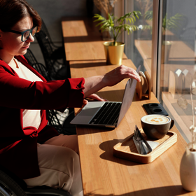 Eine Frau im Rollstuhl sitzt mit ihrem Laptop an einem Tisch. Daneben eine grosse Tasse Kaffee.