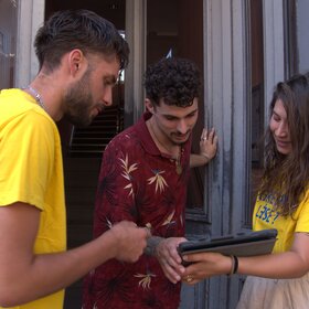 Zwei junge Menschen mit gelben T-Shirts sprechen mit einem Mann an der Haustüre.