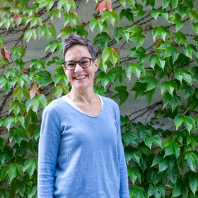 [Bitte in "Französich" uebersetzen:] Julia Eugster blickt lächelnd in die Kamera und steht vor einem grünen Hintergrund mit vielen Pflanzen.