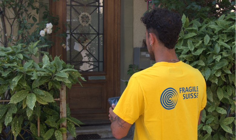 Ein junger Mann mit gelbem FRAGILE Suisse-T-Shirt steht vor einer Haustüre mit einem Tablet in den Händen.