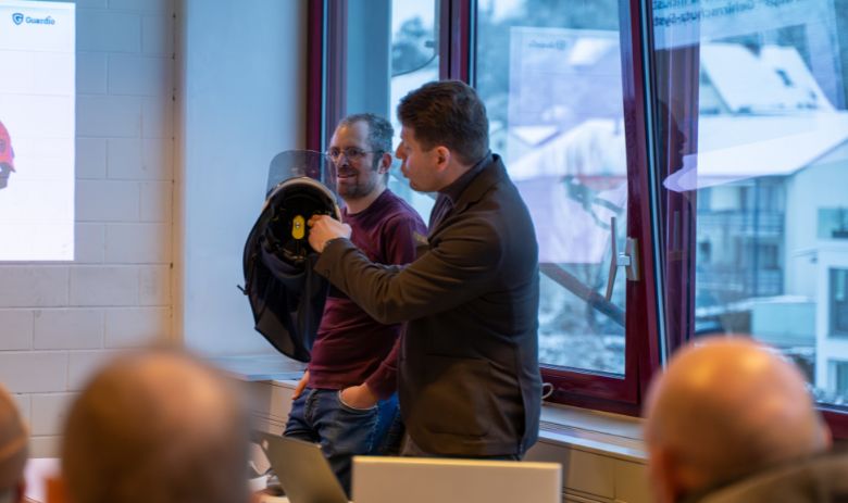 Vortrag zur Helmprävention Zwei Männer stehen vor einem Publikum. Einer der beiden Männer zeigt einen Helm.