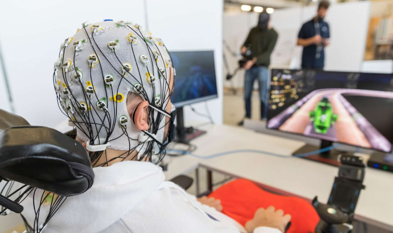 Person sitzt im Rollstuhl an einem Schreibtisch mit Monitor. Auf dem Bildschirm ist eine Art Fahrzeug auf einer Rennbahn. Die Person trägt eine Haube auf dem Kopf, aus der zahlreiche Kabel hervortreten. Auf dem Schreibtisch sind keine Steuergeräte. 