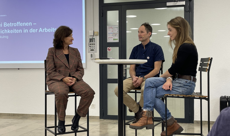 Podiumsdiskussion Fachtagung Drei Personen sitzen an einem hohen Tisch und sind im Gespräch. Im Hintergrund läuft eine Powerpoint-Präsentation.