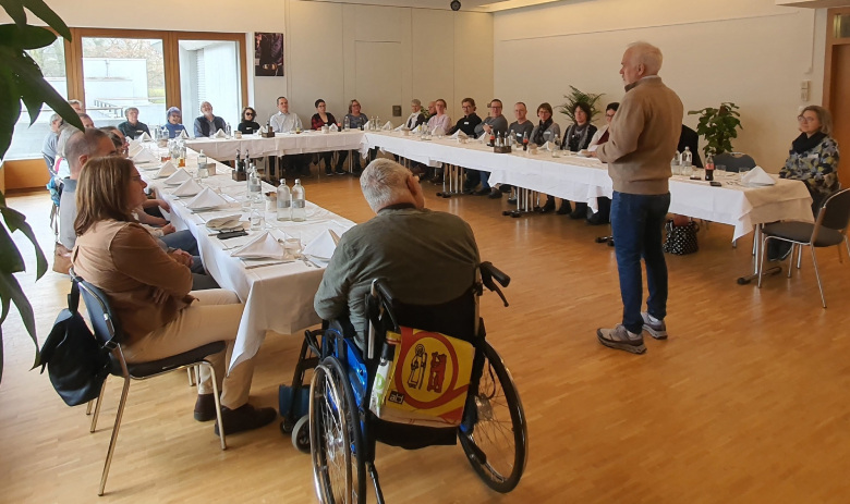 Neumitgliederanlass FRAGILE Aargau/Solothurn Ost Personen sitzen an gedeckten Tischen in einer U-Form angerichtet. In der Mitte steht eine Person und spricht zu allen.