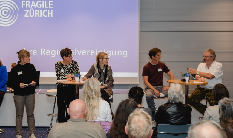 Ein Foto von hinten aus dem Publikum der Podiumsdiskussion von FRAGILE Zürich. Teilnehmende der Diskussion blicken nach links zu einem Mann im weissen Hemd.