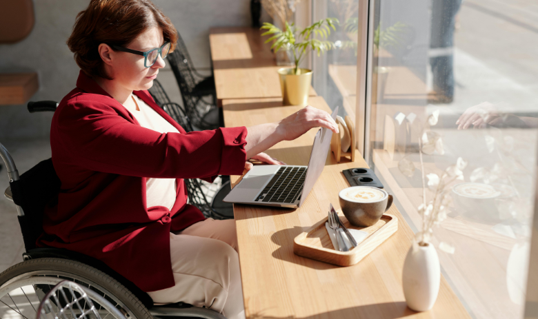 Eine Frau im Rollstuhl sitzt mit ihrem Laptop an einem Tisch. Daneben eine grosse Tasse Kaffee.