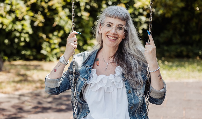 Chloé B. Chloé Buchmann Sanroma, eine modische Frau mit langen grauen Haaren sitzt auf einer Schaukel und lächelt in die Kamera.