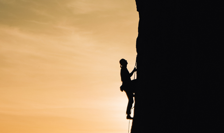 Umfrage Silhouette einer Person, die eine steile Felswand heraufklettert, vor einem Sonnenaufgang.